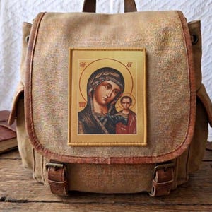 Op de afbeelding: Een beige canvas rugzak met een rechthoekige patch op de voorklep. De patch toont een religieus icoon van een vrouw en kind met gouden halo's. De rugzak is voorzien van bruine kunstlederen afwerkingen en twee gespen aan de voorzijde.