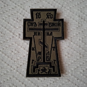 Może przedstawiać: Oliwkowa i czarna haftowana naszywka w kształcie krzyża. Naszywka zawiera tekst cyrylicą i centralny wzór krzyża. Naszywka jest prawdopodobnie emblematem religijnym lub wojskowym.