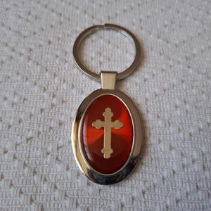 Puede incluir: Llavero de plata con un dije ovalado rojo con una cruz dorada.