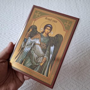 Op de afbeelding: Een gouden ingelijst icoon dat een engel met uitgespreide vleugels afbeeldt. De engel draagt een groene robe en houdt een staf vast. Het icoon heeft een rode achtergrond en is omgeven door een gouden rand.