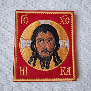 Może przedstawiać: Haftowana naszywka przedstawiająca ikonę religijną. Obraz przedstawia mężczyznę z ciemnymi włosami i brodą, na tle złotego nimbusa. Kwadratowa naszywka ma czerwone tło ze złotą obwódką i haftowanymi greckimi literami.