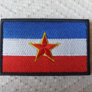 Peut inclure: Un patch brodé rouge, blanc et bleu avec une étoile rouge au centre. L'étoile a un contour jaune. Le patch a une bordure noire.