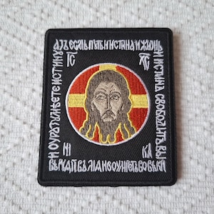 Może przedstawiać: Wyszywana kwadratowa naszywka z ikoną religijną. Centralny obraz to szczegółowe przedstawienie brodatej postaci w czerwono-żółtym okręgu. Biały tekst otacza obraz na czarnym tle.