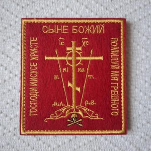 Könnte beinhalten: Ein roter gestickter Aufnäher mit goldener Stickerei, der ein Kreuz und den Text "СЫНЕ БОЖИЙ ГОСПОДИ ИИСУСЕ ХРИСТЕ ПОМИЛУЙ МЯ ГРЕШНОГО" in kyrillischer Schrift zeigt.