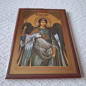 Op de afbeelding: Een religieus icoon dat een engel met vleugels afbeeldt, die een staf vasthoudt. De engel draagt een lange, vloeiende robe en heeft een halo boven zijn hoofd. Het icoon is ingelijst met goud en heeft een rode achtergrond.