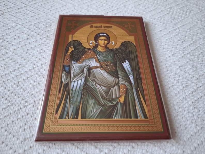 Op de afbeelding: Een religieus icoon dat een engel met vleugels afbeeldt, die een staf vasthoudt. De engel draagt een lange, vloeiende robe en heeft een halo boven zijn hoofd. Het icoon is ingelijst met goud en heeft een rode achtergrond.