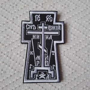 Op de afbeelding: Zwart en wit geborduurde patch in de vorm van een kruis. De patch heeft ingewikkelde details met Cyrillische tekst en een zilveren omtrek. Het kruis staat tegen een getextureerde witte achtergrond.