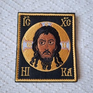 May include: Embroidered patch featuring a religious icon of a man with dark hair and a beard, set within a gold halo. The image is framed by a black border with gold lettering in Greek. The patch is square-shaped.