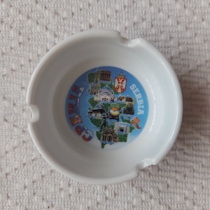 以下が含まれることがあります： 白いセラミック製の灰皿。青と白のデザインで、セルビアの地図とランドマークが描かれており、「セルビア」という文字が書かれています。