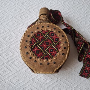 May include: A round, brown leather flask with a decorative, embroidered centre featuring red, black, and yellow geometric patterns. The flask has a braided edge, black studs, and a matching strap.