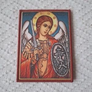 May include: Rectangular patch depicting a religious icon of an angel holding a sword and shield. The angel has red hair and a golden halo. The background is blue, and the patch is framed with a red border.