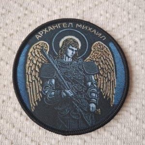 Può includere: Una toppa ricamata nera con sfondo blu che rappresenta un angelo con ali e una spada. Il testo "АРХАНГЕЛ МИХАИЛ" è scritto attorno al bordo della toppa.