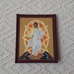 May include: A rectangular patch featuring a religious icon. The image depicts a figure in white robes with a halo, set against a gold and orange starburst design. The patch has a dark brown border.
