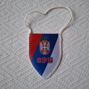 Könnte beinhalten: Ein wimpelförmiger Anhänger mit dem serbischen Flaggenmuster. Das Design zeigt die Farben Rot, Blau und Weiß. Das serbische Wappen befindet sich in der Mitte des blauen Bereichs, darunter die kyrillischen Buchstaben "СРБ".