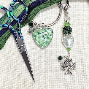 May include: A collection of craft supplies, including a pair of scissors with a blue and green paisley design, a heart-shaped charm with shamrocks, and a beaded charm with a Celtic knot. Green and blue embroidery floss is also visible.
