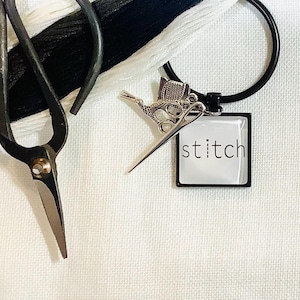 May include: A silver key chain with a small pair of scissors and a spool of thread charm. The key chain has a black ring and a square charm with the word "stitch" in white lettering.