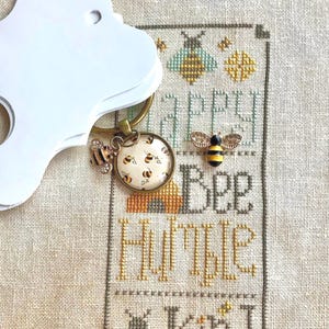 Könnte beinhalten: Ein Kreuzstichmuster mit dem Text "Bee Humble Be Kind" und einem Bienenanhänger an einem Schlüsselring. Der Anhänger zeigt eine Biene mit gelben und schwarzen Streifen und einen kleinen Bienenstock.