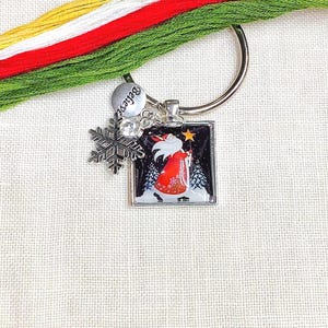 May include: A silver keyring with a Christmas-themed charm. The charm depicts Santa Claus in a snowy scene. Additional charms include a snowflake and a disc with the word "Believe". Colourful embroidery floss is visible.