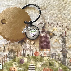 Peut inclure: Un porte-clés en métal noir avec un cabochon en verre circulaire portant l'inscription "Yes, I CAN DRIVE A stick" en noir, vert et violet, accompagné d'une petite breloque en argent représentant une sorcière sur un balai et des étiquettes dorées.