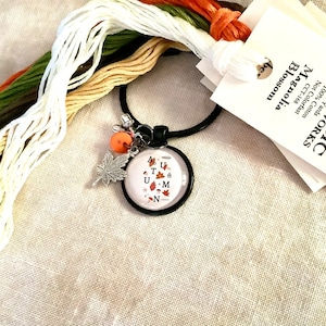 May include: A black and white charm with the word "AUTUMN" spelled out with autumn leaves. The charm is attached to a black key ring with a small orange bead and a silver leaf charm. The key ring is surrounded by spools of embroidery floss in white, beige, orange, and green.