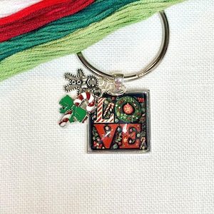 May include: A silver-toned metal keychain featuring a square pendant with the word LOVE printed in red and white, decorated with Christmas wreaths and ornaments. Two small charms, a gingerbread person and a candy cane, hang from the ring.