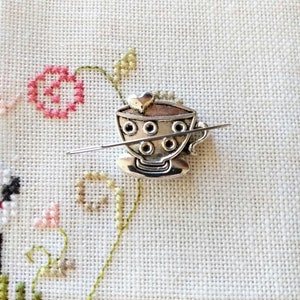 Puede incluir: Encanto de taza de t&eacute; de metal plateado con un asa en forma de coraz&oacute;n y una fila de agujeros alrededor del borde. El encanto est&aacute; enhebrado en una aguja.