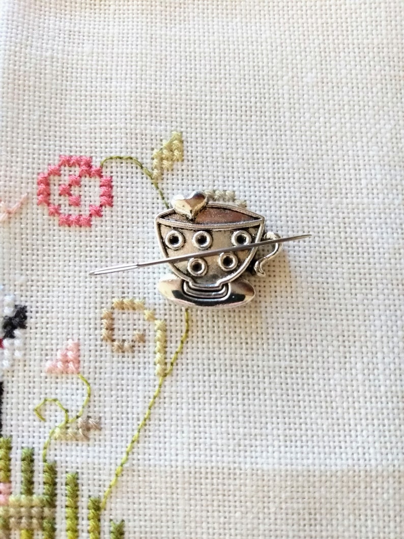 Puede incluir: Encanto de taza de t&eacute; de metal plateado con un asa en forma de coraz&oacute;n y una fila de agujeros alrededor del borde. El encanto est&aacute; enhebrado en una aguja.