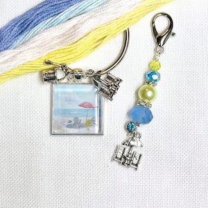 May include: A silver-toned charm with a beach scene, featuring a red and white striped umbrella, beach chairs, and ocean view. A beaded charm with blue and yellow beads and a silver clasp is also present. Embroidery floss in shades of blue, white, and yellow is in the background.