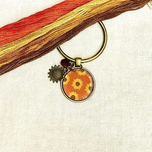 May include: A gold-colored metal keyring with a round pendant featuring a sunflower pattern on an orange background. A small sunflower charm and a dark red bead dangle from the keyring. Strands of orange, yellow, and brown embroidery floss are in the background.