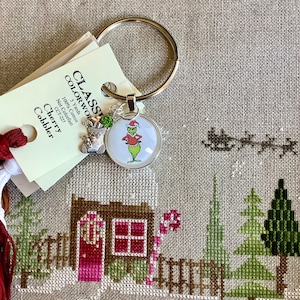 May include: A silver key chain with a round charm featuring a cartoon Grinch wearing a Santa hat. The charm is attached to a silver ring. The key chain is displayed on a beige fabric background with a cross-stitch design of a small house with a red door and a candy cane.