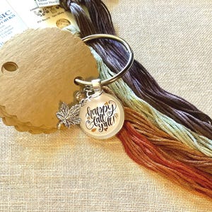 May include: A silver charm with a round glass cabochon featuring the text "Happy Fall Y'all!" in brown lettering. The charm is attached to a silver ring with a small silver leaf charm. The charm is surrounded by a variety of colourful embroidery floss.