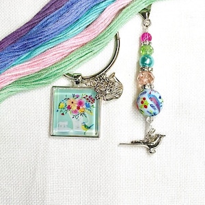 Puede incluir: Un imán para agujas plateado con un colgante cuadrado con motivos florales y un charm de cuentas. El colgante presenta un jarrón de flores y un pajarito. El charm tiene cuentas de colores y un charm de pájaro. También se ven hilos de bordar en colores pastel.