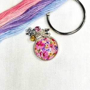 Peut inclure: Un pendentif rond en argent avec un motif floral rose, violet et jaune. Le pendentif est orné de petites pierres précieuses colorées et d'une fleur argentée. Un paquet de fil à broder rose, violet et bleu et un cercle en argent sont également dans l'image.