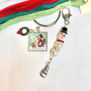May include: A silver-toned keychain set featuring a square pendant with a snowman and reindeer illustration, a dangling snowman charm made of white beads, a black hat, and red rhinestones, accompanied by white, red, and green embroidery floss.