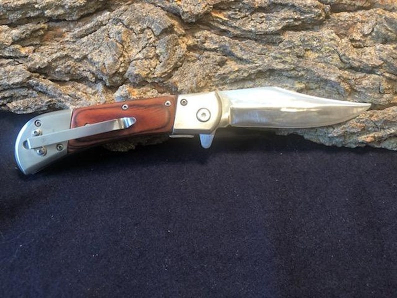 May include: A silver folding knife with a wood handle. The blade is closed and the knife is resting on a piece of bark.