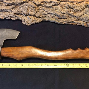May include: A hand-forged axe with a wooden handle. The axe head has a hammered finish and the handle is made of dark wood with a textured finish. The axe is 17 inches long.