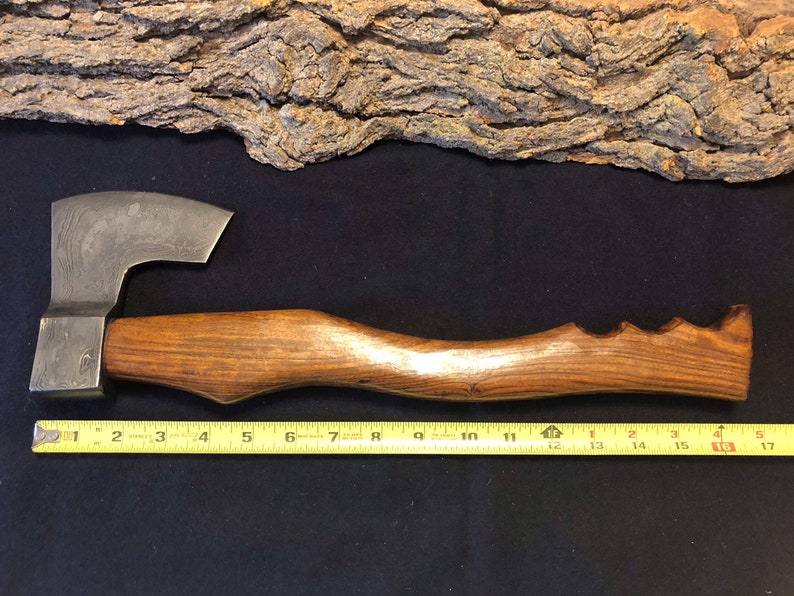 May include: A hand-forged axe with a wooden handle. The axe head has a hammered finish and the handle is made of dark wood with a textured finish. The axe is 17 inches long.