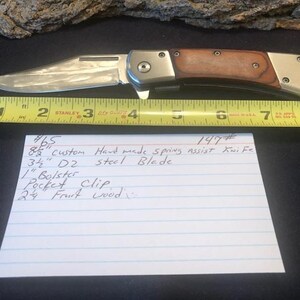 May include: A close-up of a silver folding knife with a wood handle. The knife is 3 1/2 inches long and is made of D2 steel. The knife has a spring assist and a pocket clip. The knife is labeled #147 and #465.