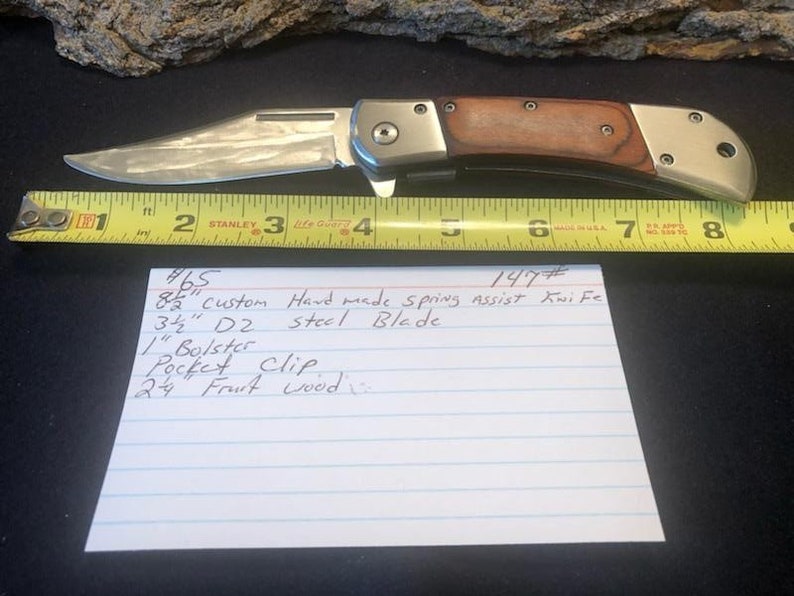 May include: A close-up of a silver folding knife with a wood handle. The knife is 3 1/2 inches long and is made of D2 steel. The knife has a spring assist and a pocket clip. The knife is labeled #147 and #465.