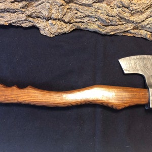 May include: A hand-forged axe with a wooden handle. The axe head has a hammered finish and a slight curve to the blade. The handle is made of dark wood with a natural grain pattern.
