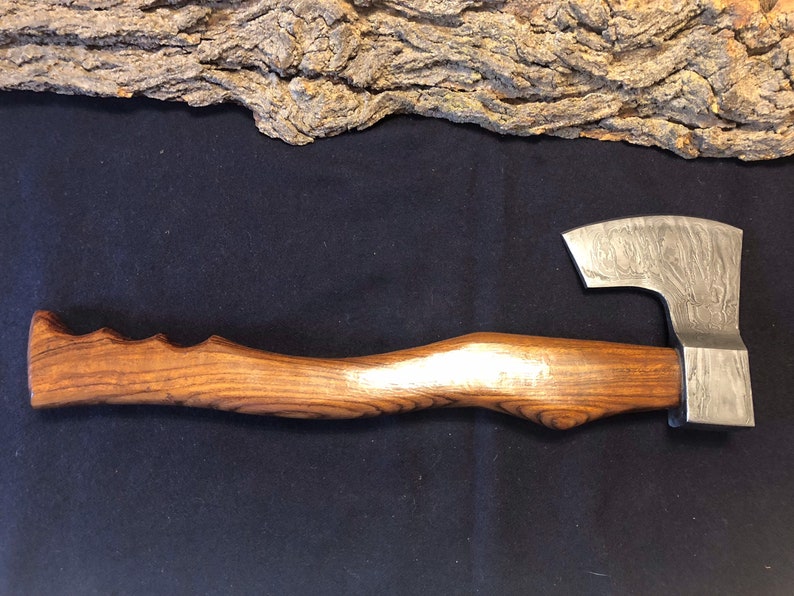 May include: A hand-forged axe with a wooden handle. The axe head has a hammered finish and a slight curve to the blade. The handle is made of dark wood with a natural grain pattern.