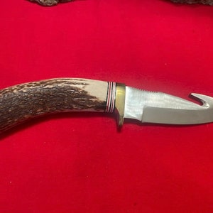 May include: A hunting knife with a brown and white antler handle and a silver blade with a hook at the end.