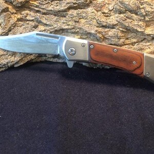 May include: A silver folding knife with a wooden handle. The handle is a dark brown wood with a reddish hue. The blade is closed and the knife is resting on a piece of bark.