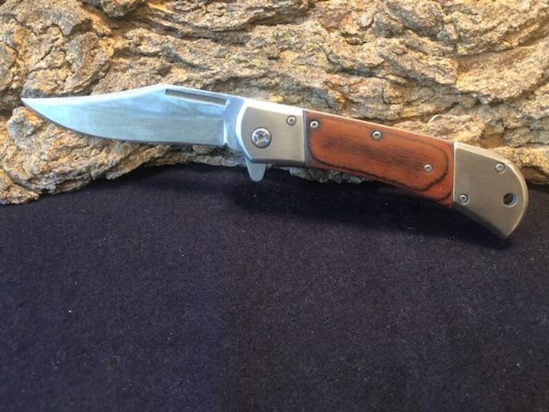May include: A silver folding knife with a wooden handle. The handle is a dark brown wood with a reddish hue. The blade is closed and the knife is resting on a piece of bark.