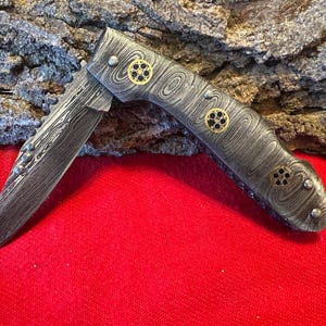 May include: A silver pocket knife with a patterned handle and gold accents. The blade is partially open and has a serrated edge.