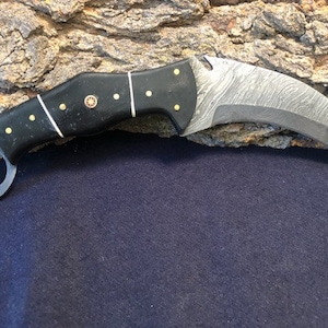 May include: A black and silver karambit knife with a black handle and a ring on the end of the blade.