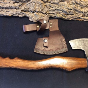 May include: A hand-forged axe with a wooden handle and a leather sheath. The axe head has a Damascus steel pattern and the handle is made of wood with a natural grain. The sheath is brown leather with a decorative stitching pattern.
