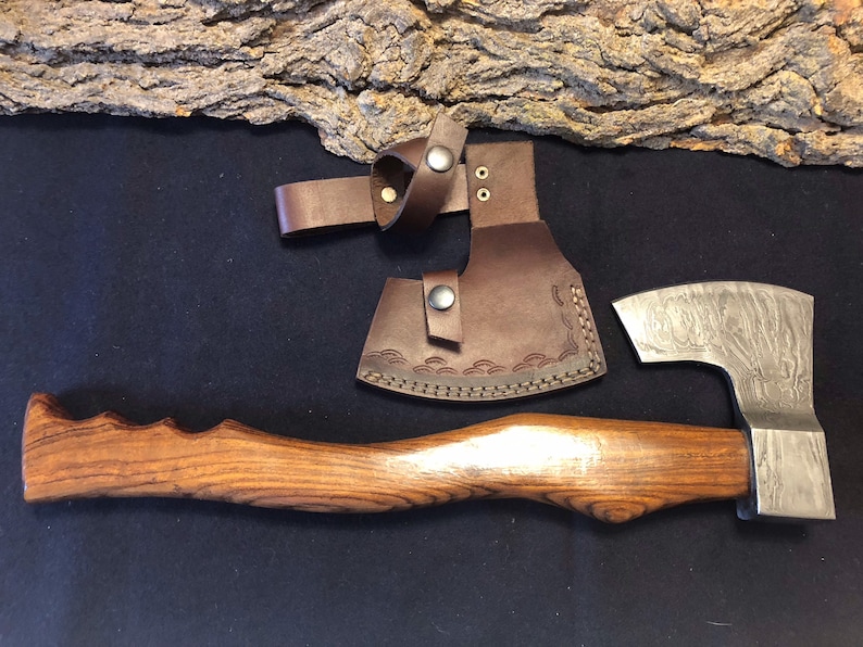 May include: A hand-forged axe with a wooden handle and a leather sheath. The axe head has a Damascus steel pattern and the handle is made of wood with a natural grain. The sheath is brown leather with a decorative stitching pattern.