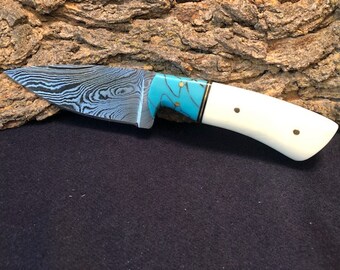May include: A handmade knife with a blue and white Damascus steel blade and a white bone handle with turquoise accents.