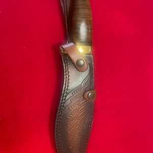 May include: A brown leather knife sheath with a decorative embossed design. The sheath has a brass snap closure and a brass tip.